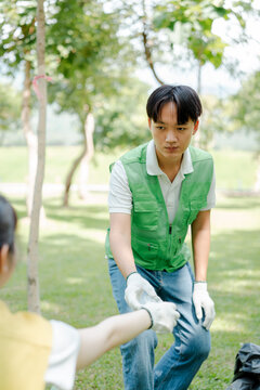 Young asian man in a green vest collecting plastic waste into a black bag outdoors, accepting more trash from another person while cleaning a park and promoting environmental responsibility
