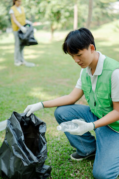 Young asian man volunteering for environmental cleanup. Collecting plastic waste in a black trash bag from green grass. With another volunteer in background