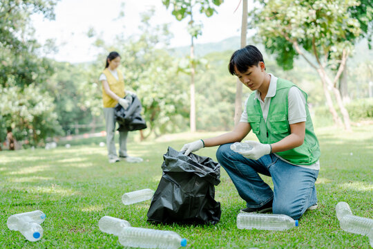 Young volunteers in vests and gloves collect plastic bottles and trash into black bags on park grass, working together to clean the green space and protect the environment