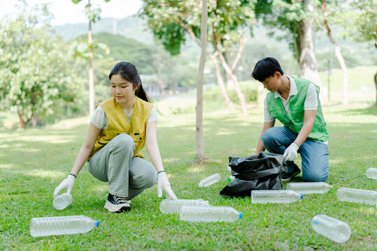 Young asian volunteers cleaning up nature, collecting plastic waste bottles from green grass in a public park, focusing on environmental protection and community recycling initiative