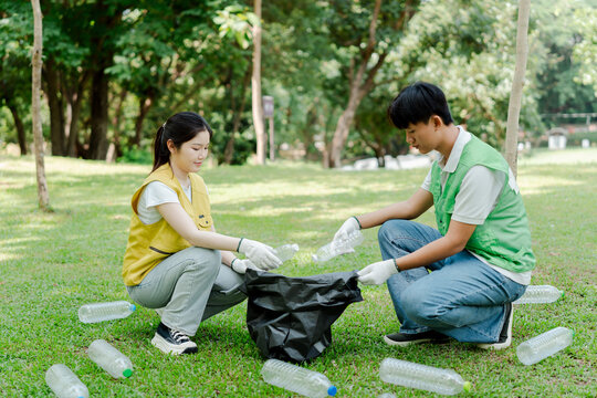Young asian man and woman volunteers cleaning up park, collecting plastic bottles into a trash bag, promoting environmental protection and waste recycling