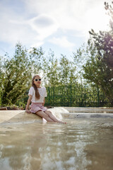 Fototapeta premium A stylish girl wearing sunglasses, a white T-shirt and a pink skirt sits at the edge of a shallow pool on a sunny summer day. She relaxes outdoors in a green park, enjoying warm weather
