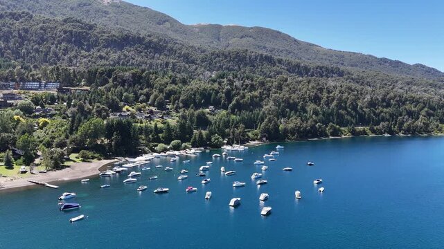 Stunning aerial perspective of numerous white boats and yachts moored in a tranquil blue bay of Nahuel Huapi Lake in Patagonia, Argentina. Lush alpine pine forests cover the rolling mountainside.