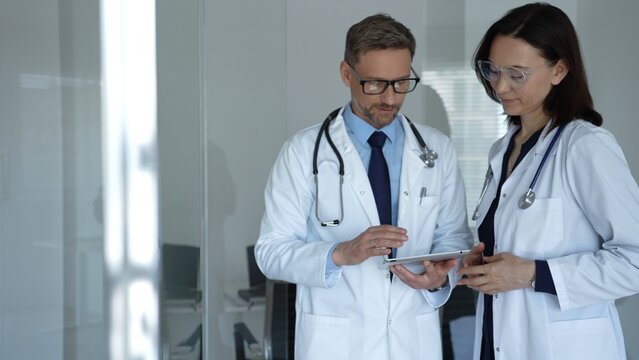 Male and female doctors collaborate over patient data on a tablet in a clinic, both in lab coats with stethoscopes, using digital tech for modern patient care and diagnosis