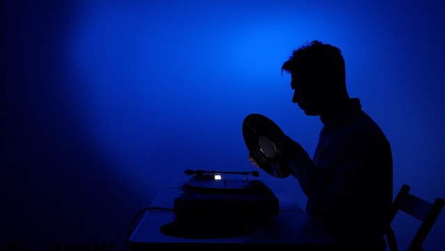 Man in silhouette holding and placing an lp record on a vintage turntable in a dark room, illuminated by a dramatic blue light, highlighting classic audio technology and musical nostalgia