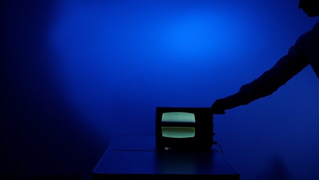 Silhouette arm adjusting an old cathode ray tube television set, displaying a green screen against a vibrant blue background, symbolizing vintage technology and past media