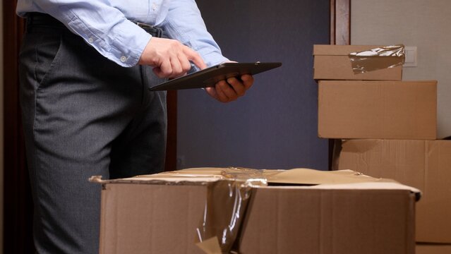 Deliveryman using a digital tablet to manage incoming packages and update logistics information, standing next to a stack of cardboard boxes for efficient shipping and e commerce fulfillment