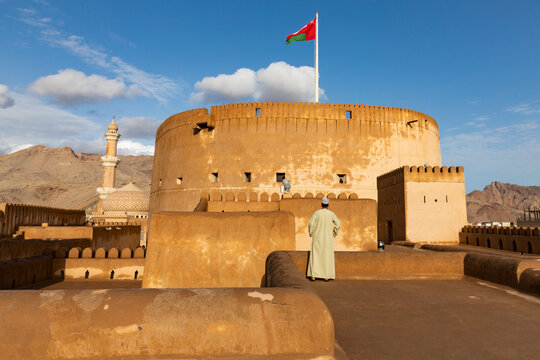Nizwa Fort with Omani flag and man in traditional dress under warm light, showcasing heritage, culture, and national identity. Nizwa, Oman - April 6 2026