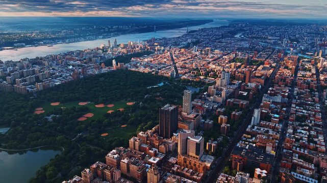Overhead drone pan across northern Central Park, Manhattan. Twilight sky over ballfields, Harlem rooftops and distant Hudson River bridge on the horizon.
