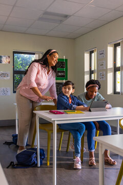 Teacher with two children leaning over table in classroom, gesturing toward tablet and notebooks