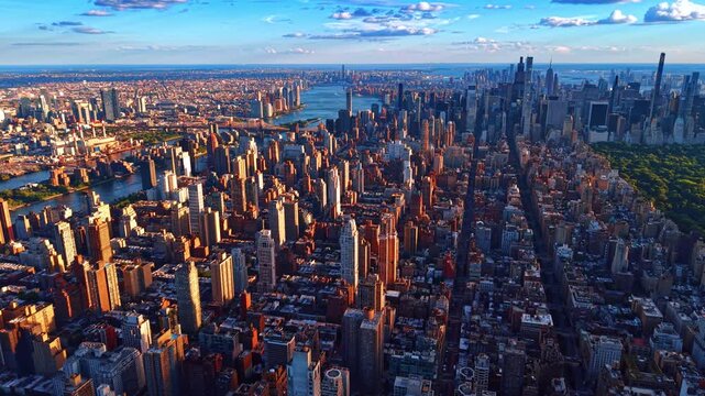 Dense Manhattan skyline stretching toward East River at golden hour. Elevated drone view over brownstones, avenues and midtown towers under blue sky with clouds.