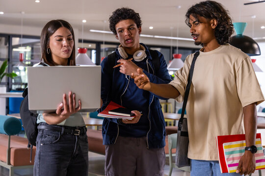 Diverse college students studying in study lounge around silver laptop, holding notebooks, pointing