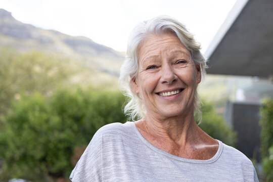 Senior woman smiling outside near building overhang, wearing light gray knit top and stud earrings