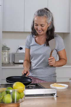Middle-aged woman standing at stove preparing food with nonstick pan and spatula in kitchen