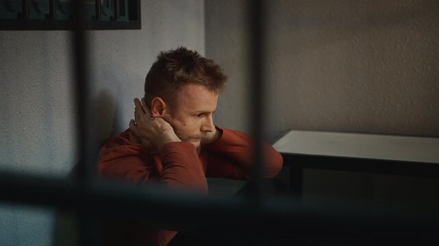 Unhappy young jailed man sitting on the floor behind bars, feeling desperate