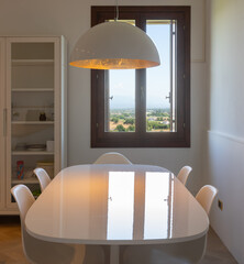 Obraz premium Front view of an oval white table, with chairs underneath, a sideboard to the left, and a window overlooking the Italian hills. Country house