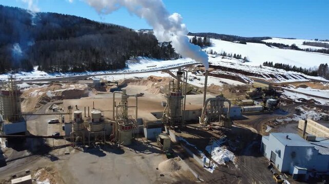 Drone footage flying over a snowy sawmill, passing through steam from a towering chimney and revealing cyclones, baghouses, and wood processing facilities.