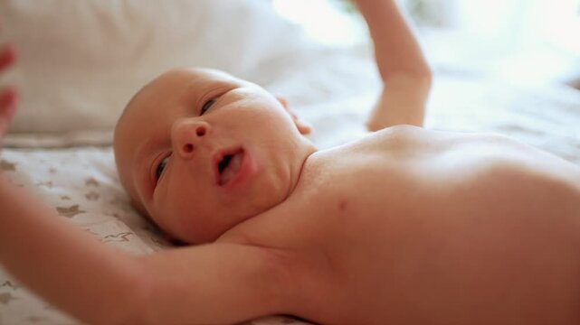 Cute newborn baby girl with blue eyes lying on her back on a bed. The infant is yawning, stretching, and moving her tiny arms and hands in a sunlit room, embodying pure innocence