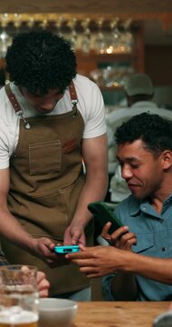 Man, waiter and customer with phone on pos machine for payment, table bill or service in local pub. Male person, group or payer with smartphone tap for free drinks, tab or treating friends in bar