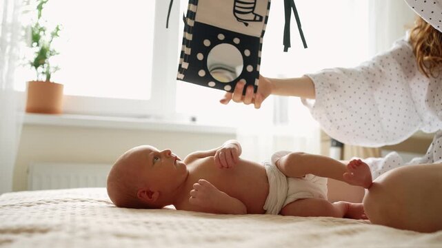 Curious newborn baby in a diaper lying on a bed while its mother shows high contrast black and white pictures, promoting early visual sensory stimulation and brain development