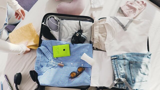 Overhead view of hands packing clothes and essentials into a travel suitcase. Summer vacation preparation and luggage packing for tourism. High quality 4k footage