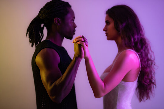 Multiracial couple holding hands and looking at each other in studio against lilac background