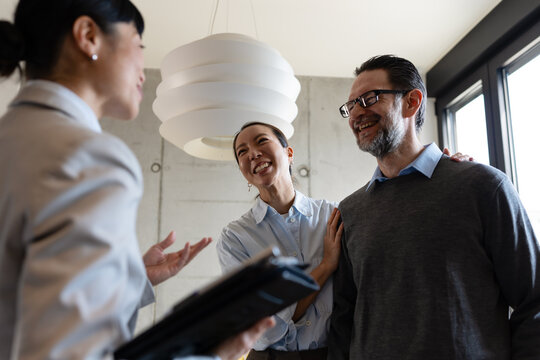 Happy diverse couple viewing a modern apartment with a professional real estate agent. Smiling multicultural clients discussing property buying, renting, or investing during a house tour.