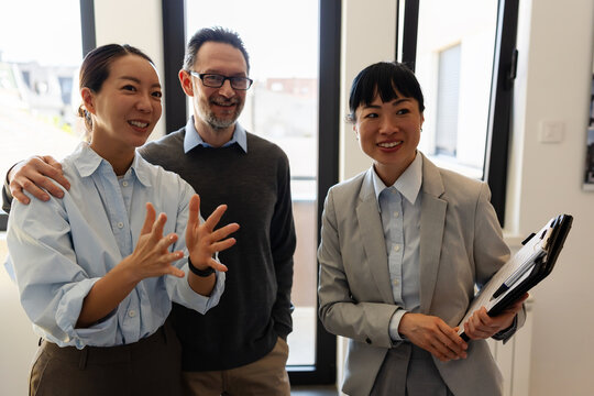 Happy diverse couple viewing a modern apartment with a professional real estate agent. Smiling multicultural clients discussing property buying, renting, or investing during a house tour.