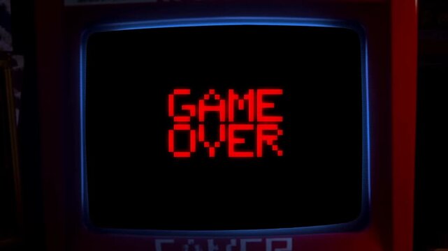 Static shot of arcade cabinet crt screen displaying red pixelated game over text animating and flickering in dark retro arcade interior