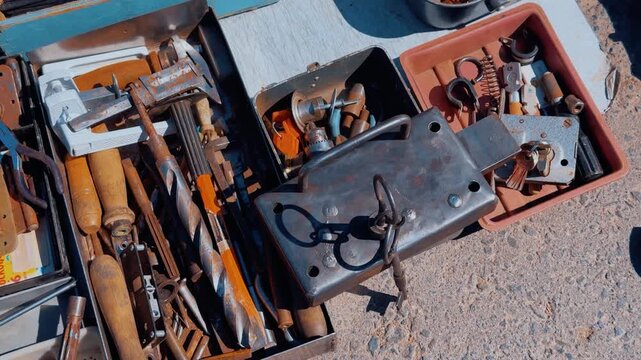 Old rusty tools in boxes laid on sunlit asphalt at busy flea market, assorted hammers, chisels, pliers, scissors and metal parts
