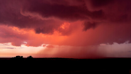 Obraz premium Spectacular Crimson Thundercloud Formation Illuminated by Sunset Above a Dark Countryside Horizon