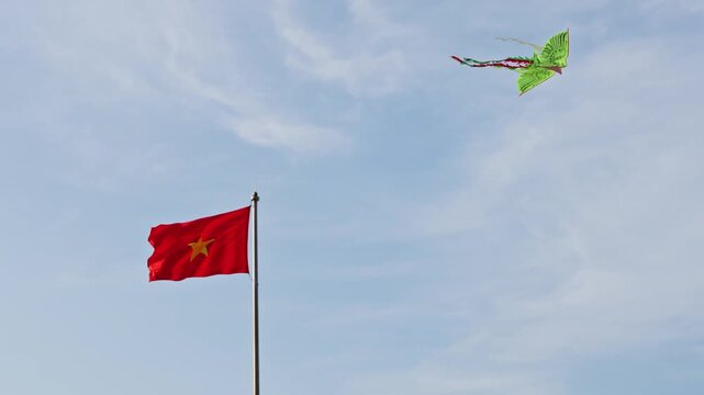 Vietnamese flag waving against a blue sky. A green kite drifts in the upper sky while a red Vietnamese flag waves from a tall pole below, set against a bright blue daytime background with light clouds