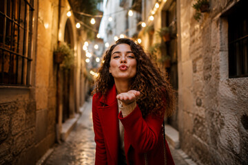 Obraz premium Young Woman Blowing Kiss In Charming Historic European Street With Warm Lights Romantic Travel Lifestyle Portrait Photography Concept