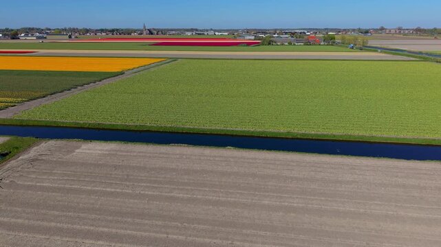 Aerial view of multicolored agricultural fields with plowed soil, green crops yellow and red flower bands water canals rural town church steeple and expansive farmland under clear daylight conditions