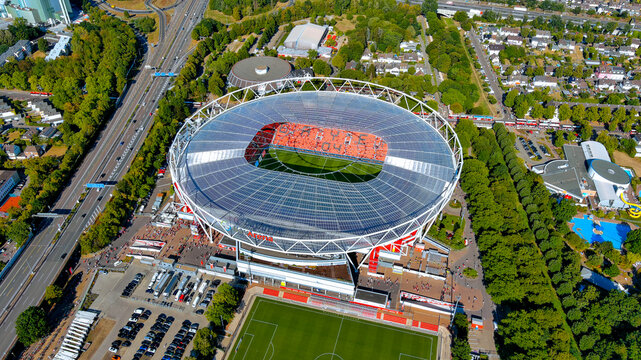 Aerial drone view of BayArena football stadium in Leverkusen Germany on a sunny day featuring the circular glass roof green pitch and surrounding road infrastructure.
