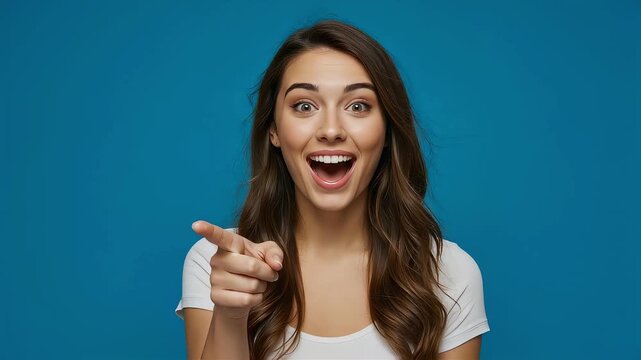 A woman points to the camera with an expression of surprise and joy in front of a blue background.