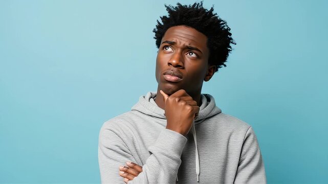 A young Black man stands with his hand on his chin, looking confused and deep in thought, in front of a light blue background.