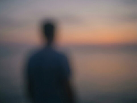 Silhouette of a person standing by the water's edge at sunset, with soft colors blending in the sky and a blurred background. Loneliness and depression concept, space for text