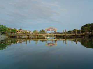 Fototapeta premium Historic bridge reflected in calm water along a peaceful canal in Hoi An Ancient Town.