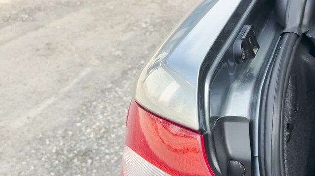 An open silver car trunk door reveals the inner latch next to a gravel path. Closing it creates a loud clicking and deep thudding sound.