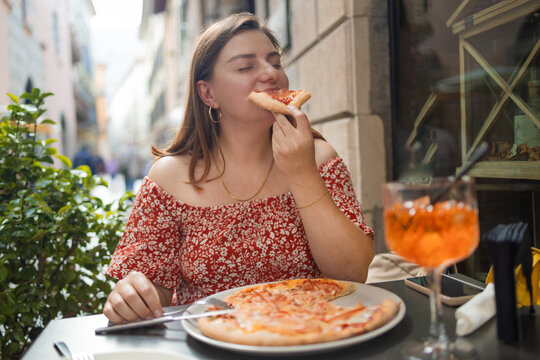 smiling beautiful young woman enjoying a slice of fresh Italian pizza at a cozy outdoor restaurant table. She is having lunch with a traditional orange cocktail on a narrow picturesque European street