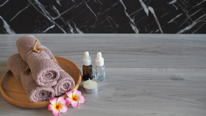 Luxury spa still life with essential oil dropper bottles (amber and clear glass), a scented candle, soft pink mauve towels tied with rope, and pink frangipani flowers on a light gray wooden table. Pre © kanjana