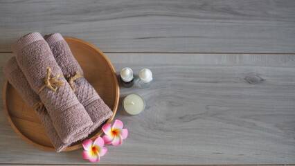 Luxury spa still life with essential oil dropper bottles (amber and clear glass), a scented candle, soft pink mauve towels tied with rope, and pink frangipani flowers on a light gray wooden table. Pre © kanjana