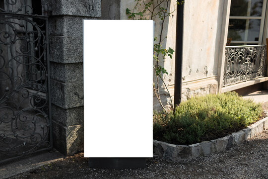 Close-up of an empty white informational board or standing signpost on a stone walkway next to a garden bed outdoors.