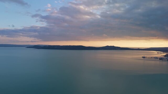 In Tihany, Lake Balaton stretches toward the horizon with calm water reflecting a cloudy sunset sky, bordered by hills and a quiet shoreline seen from a drone perspective.
