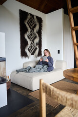 Obraz premium Woman reading book on sofa wrapped in blanket, enjoying cozy home interior with warm textures and fireplace, calm leisure time and relaxing lifestyle concept