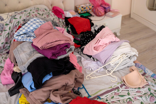 A slightly elevated angle view of a bedroom shows two distinct large clothing piles on a bed covered in floral bedding, with white plastic hangers scattered between them and a beige bra and black shoe