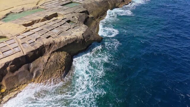 Aerial view of the Xwejni salt pans in Żebbuġ, Gozo, where geometric salt basins edge the rocky coast above clear blue Mediterranean waters and foaming sea waves.