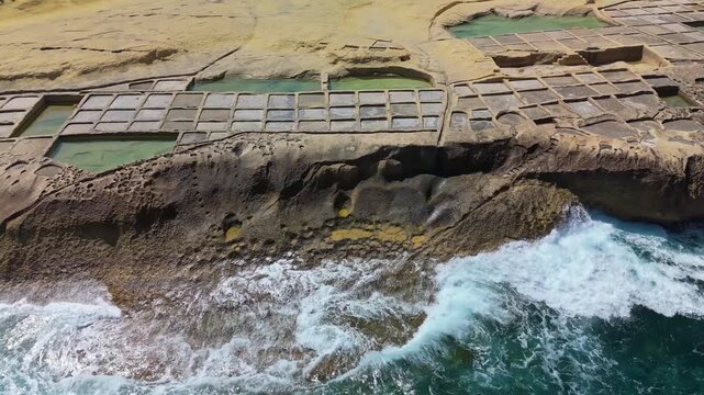 Xwejni salt pans in Żebbuġ, Gozo line the rocky Maltese coastline beside turquoise Mediterranean waves, forming geometric patterns from a drone perspective.