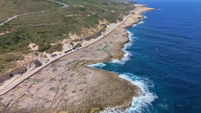Xwejni&rsquo;s centuries-old salt pans unfold along the rugged coast of Żebbuġ, Gozo, where geometric pools meet vivid Mediterranean blues in a sweeping summer drone view.